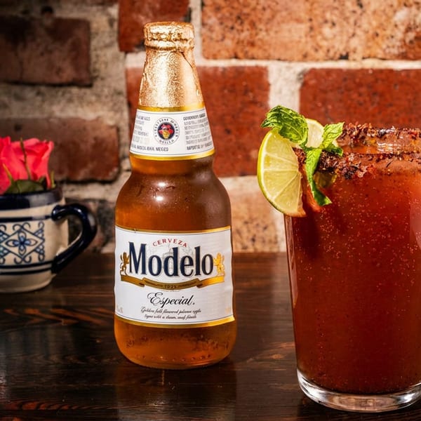 Modelo Especial beer bottle next to a michelada cocktail with lime and mint, on a wooden table.