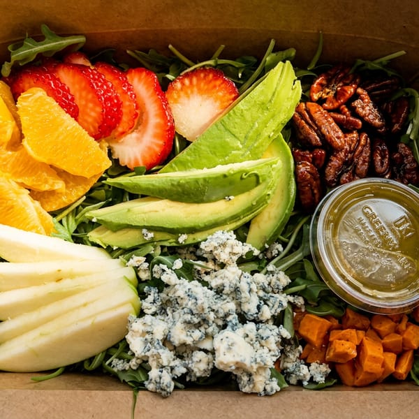 Colorful salad in a cardboard box with strawberries, oranges, avocado, pecans, blue cheese, and sweet potatoes.