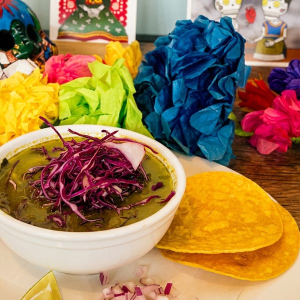 Green pozole with shredded red cabbage, radishes, and tortillas, surrounded by colorful paper flowers.