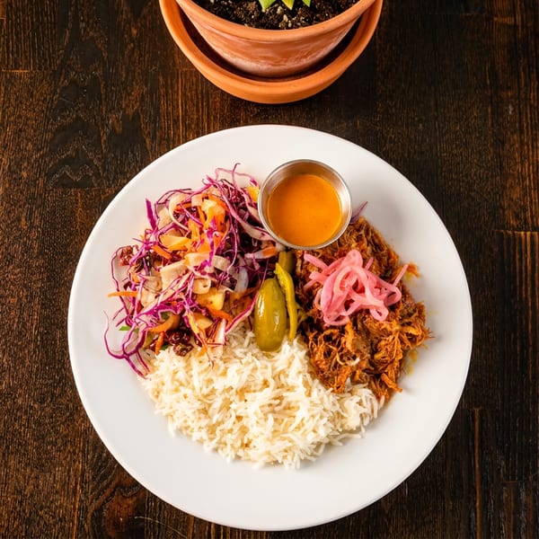 A white plate with rice, shredded meat, pickled onions, salad, and a side of sauce.