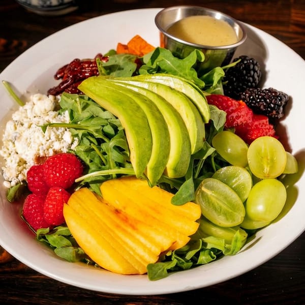 A vibrant salad with avocado, berries, grapes, mango, feta, and dressing in a white bowl.