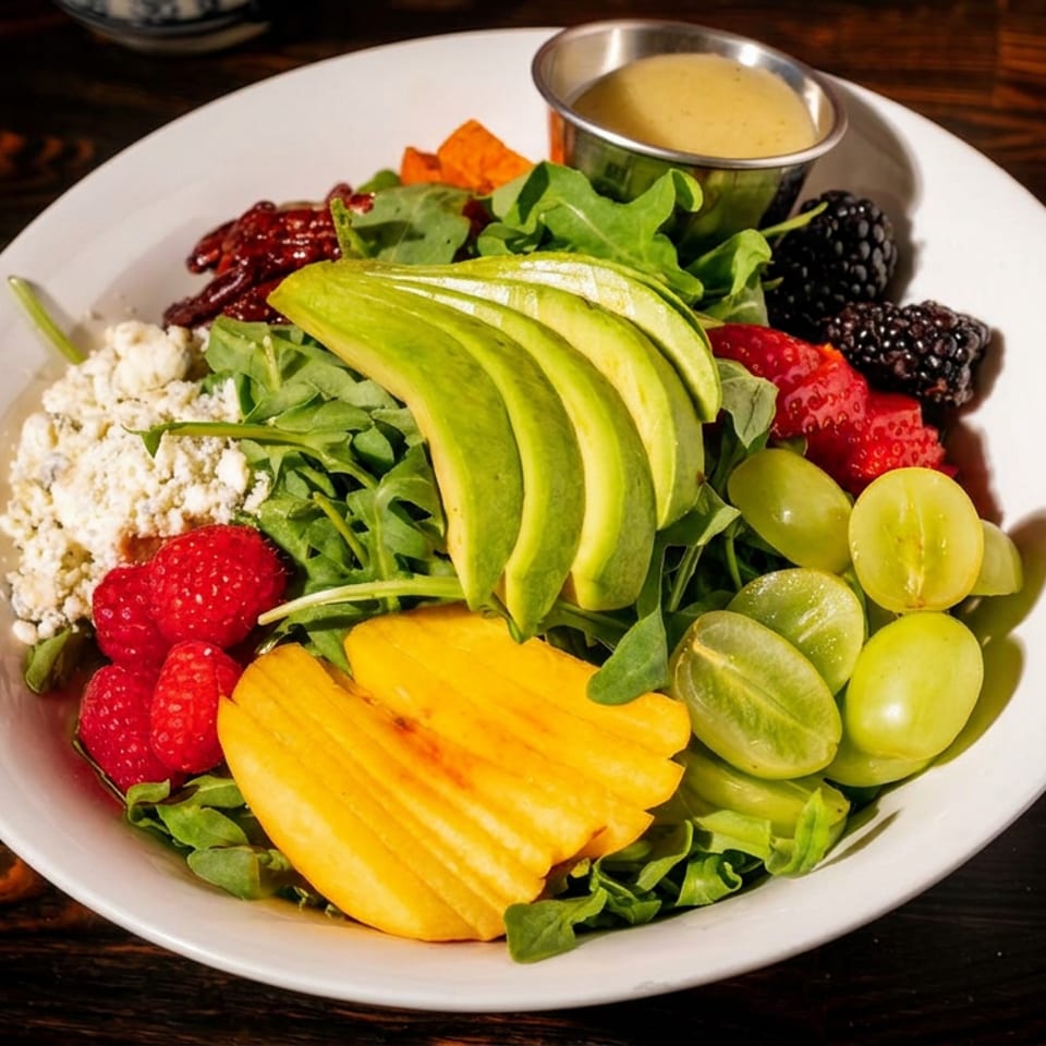 A vibrant salad with avocado, berries, grapes, mango, feta, and dressing in a white bowl.