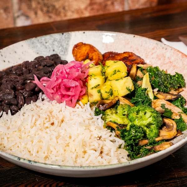 A colorful bowl of rice, black beans, pickled onions, pineapple, plantains, broccoli, and kale.