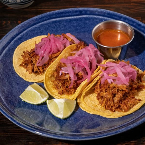 Three carnitas tacos with pickled red onions, lime wedges, and a side of red sauce on a blue plate.